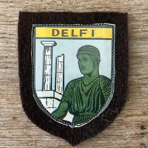 Op de afbeelding: Een vintage patch met het woord "DELFI" in geel, met een afbeelding van een groene figuur en klassieke zuilen. De schildvormige patch heeft een bruine viltrug.
