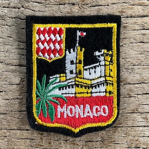 Op de afbeelding: Geborduurde patch met het wapen van Monaco. Het ontwerp omvat een schild met een rood-wit geblokt patroon, een kasteel, een palmboom en het woord "MONACO" in witte letters op een rode achtergrond, alles omlijnd in geel.