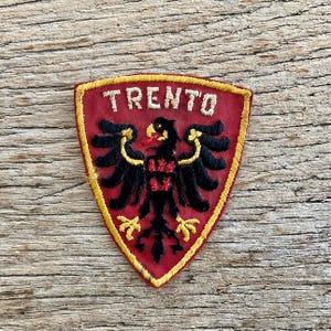 May include: A red, shield-shaped patch with the word "TRENTO" in white lettering. The patch features a black eagle with yellow accents. The patch is bordered with yellow stitching.