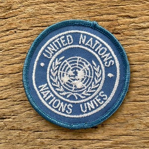 Op de afbeelding: Een ronde, blauwe patch met witte geborduurde tekst en een centraal embleem. De tekst luidt "UNITED NATIONS" en "NATIONS UNIES" rond het embleem, dat een wereldkaart omgeven door olijftakken afbeeldt.