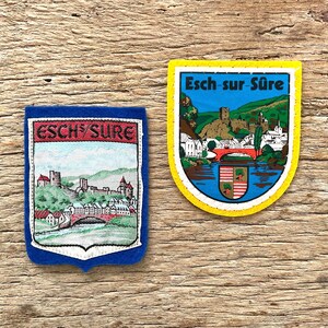 Op de afbeelding: Twee geborduurde patches met de stad Esch-sur-Sûre. De linker patch heeft een blauwe rand en toont een kasteel en een brug. De rechter patch heeft een gele rand, de tekst Esch-sur-Sûre en een kleurrijke illustratie van de stad.