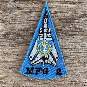 Peut inclure: Écusson bleu triangulaire avec un avion à réaction stylisé blanc et noir et un symbole de cible jaune et blanc. Le texte "MFG 2" est imprimé en blanc. Bordure bleue. Il s'agit probablement d'un emblème militaire ou aéronautique.