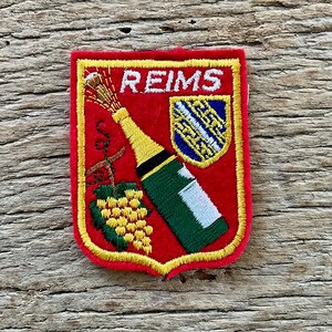 Könnte beinhalten: Ein schildförmiger, bestickter Aufnäher mit rotem Hintergrund und gelbem Rand. Er zeigt das Wort REIMS in Weiß, eine grüne Champagnerflasche mit goldenem Hals, eine gelbe Weintraube und ein kleines blaues Schild mit weißem Muster.