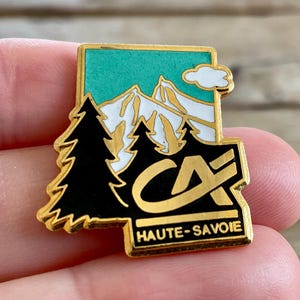 Op de afbeelding: Een goudkleurige emaille pin met een berglandschap met witte toppen, een turquoise lucht en zwarte dennenbomen. De pin heeft de tekst "CA HAUTE-SAVOIE" in gouden letters.