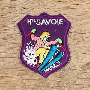 Op de afbeelding: Een paarse patch met de tekst "Hte Savoie" en een skiër. De skiër draagt een roze jas, gele broek en blauwe ski's. De patch heeft een witte rand en ligt op een houten oppervlak.