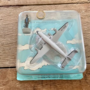 K&ouml;nnte beinhalten: Ein Vintage-Spielzeug-Flugzeugmodell in einer transparenten Kunststoffverpackung. Das wei&szlig;-silberne Flugzeug hat die Aufschrift VT-DAO auf der Seite. Eine kleine Figur und eine M&uuml;nze sind ebenfalls sichtbar. Die Verpackung hat ein blaues Himmel- und Wolkenmuster.