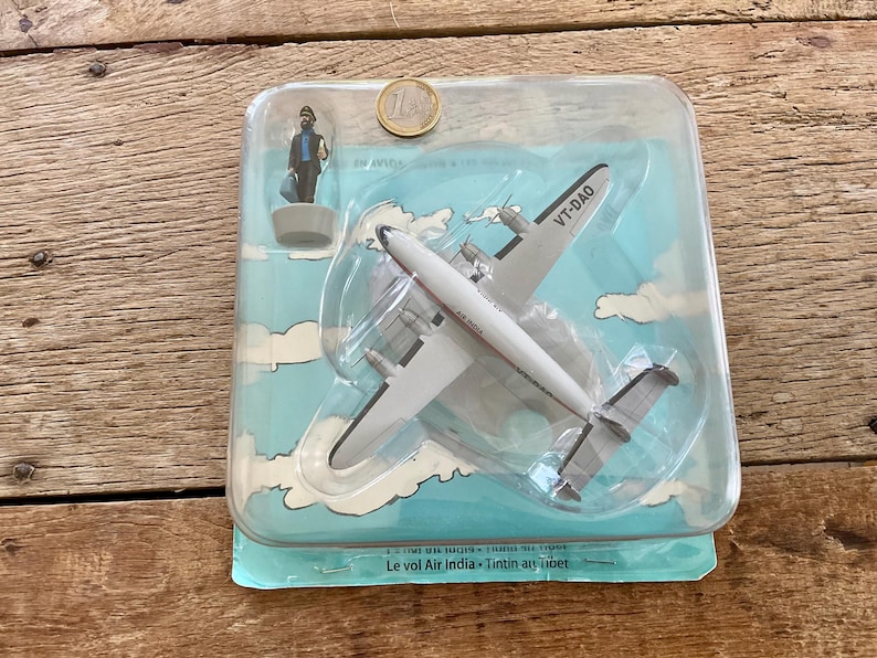 K&ouml;nnte beinhalten: Ein Vintage-Spielzeug-Flugzeugmodell in einer transparenten Kunststoffverpackung. Das wei&szlig;-silberne Flugzeug hat die Aufschrift VT-DAO auf der Seite. Eine kleine Figur und eine M&uuml;nze sind ebenfalls sichtbar. Die Verpackung hat ein blaues Himmel- und Wolkenmuster.