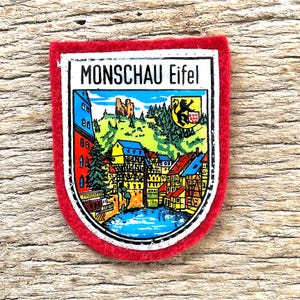Könnte beinhalten: Ein rechteckiger Aufnäher mit rotem Rand und weißem Innenraum. Der Aufnäher zeigt den Text "MONSCHAU Eifel" über einer farbenfrohen Illustration einer Stadt mit Fluss, Gebäuden und Burg. Rechts befindet sich ein schwarz-gelbes Emblem.