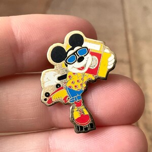 Op de afbeelding: Een metalen emaille speldje met een cartoonmuis die een blauwe 3D-bril, een geel overhemd met rode sterpatronen en een blauwe korte broek draagt. Het personage staat op rolschaatsen en draagt een rood-gele gettoblaster op de schouder.