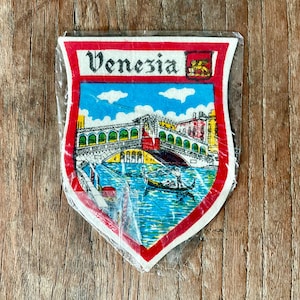 Op de afbeelding: Een schildvormige patch met een kleurrijke illustratie van Venetië, Italië. Het ontwerp omvat de Rialtobrug, gondels en het woord "Venezia" bovenaan. De patch is omlijst met een rode rand en is ingekapseld in helder plastic.