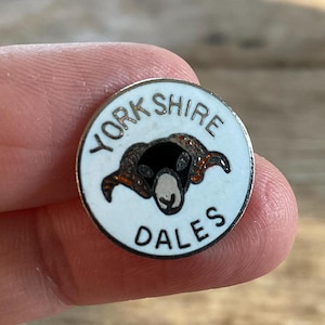 Op de afbeelding: Een ronde emaille speld met een witte achtergrond en de zwarte tekst YORKSHIRE DALES in een boog rond een centrale afbeelding van een schapenkop. Het schaap heeft een zwart gezicht, een witte snuit en bruine hoorns, gevat in een zilverkleurige rand.