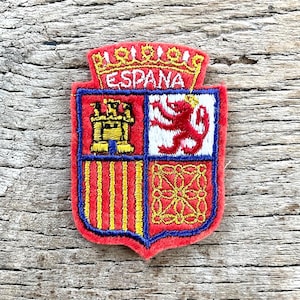 Op de afbeelding: Geborduurde patch met het Spaanse wapen. De rode patch heeft een blauwe rand en toont een gouden kroon, het woord "ESPANA", een kasteel, een leeuw en verticale strepen. Op een houten oppervlak.