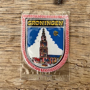 Op de afbeelding: Een patch met het woord "GRONINGEN" boven een kleurrijke illustratie van een hoge kerktoren en gebouwen tegen een blauwe lucht. De patch zit in een doorzichtige plastic hoes met een rode en witte rand.