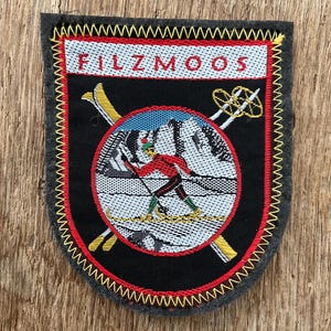 Op de afbeelding: Een zwarte patch met het woord "FILZMOOS" bovenaan. De patch toont een skiër in rood en groen, die een besneeuwde berg afdaalt, met gele ski's en stokken. De patch is omrand met rode en gele stiksels.