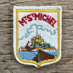 Könnte beinhalten: Gestickter Aufnäher mit dem Schriftzug "MTS MICHEL" in Rot. Der Aufnäher zeigt ein Schloss auf einer Insel mit blauem Wasser und einer braunen Brücke. Der Aufnäher hat einen goldenen Rand. Das Design ist vor einem weißen Hintergrund.
