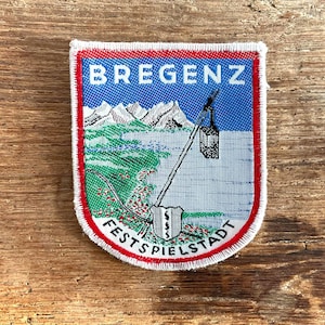 May include: Embroidered patch with the word "BREGENZ" at the top, depicting mountains, a lake, and a cable car. The text "FESTSPIELSTADT" is at the bottom. The patch has a red border. The background is a wooden surface.