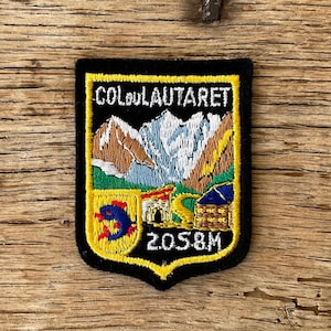 Op de afbeelding: Zwarte geborduurde patch met de tekst "COL DU LAUTARET" en "2058M". De patch toont een berglandschap met een blauwe lucht, bruine en witte bergen en een gele rand. Een schild met een rood en blauw ontwerp staat linksonder.