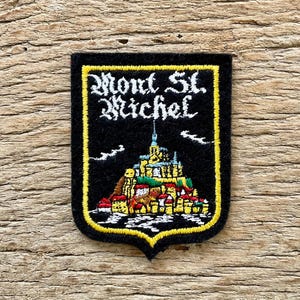 May include: Black embroidered patch with the words "Mont St. Michel" in white script above a colourful illustration of the historic island. The patch has a yellow border and a unique shield shape.