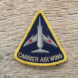 Op de afbeelding: Een marineblauwe patch met een gele rand, met een zilverkleurig vliegtuigontwerp en rode sterren op de vleugels. De tekst "CARRIER AIR WING" is in het wit geborduurd. De patch is driehoekig van vorm.