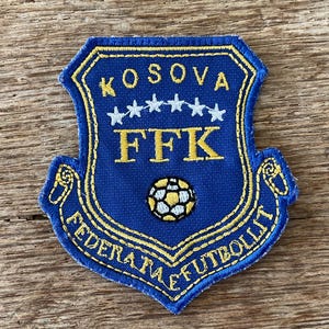 Op de afbeelding: Een blauw en geel geborduurd embleem met de tekst "KOSOVA" boven vijf witte sterren en "FFK". Het embleem bevat ook een voetbalafbeelding en de tekst "FEDERATA E FUTBOLLIT" langs de onderrand.