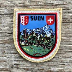 Op de afbeelding: Een Suen-patch met een berglandschapontwerp. De patch heeft een blauwe lucht, besneeuwde bergen en een dorp. Het woord "SUEN" staat bovenaan gedrukt, met een rood-wit Zwitsers kruis rechts.
