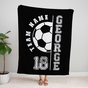 May include: Black blanket with a white soccer ball graphic, the text "TEAM NAME" and the name "GEORGE" in a vertical column. The number "18" is also displayed. The blanket is hanging in front of a wall.