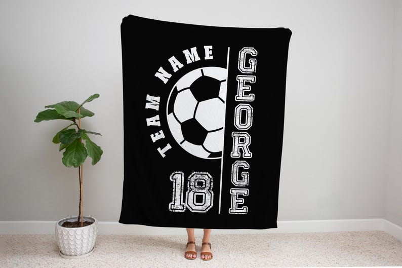 May include: Black blanket with a white soccer ball graphic, the text "TEAM NAME" and the name "GEORGE" in a vertical column. The number "18" is also displayed. The blanket is hanging in front of a wall.
