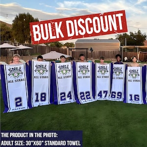 Puede incluir: Un grupo de personas sostienen toallas blancas y azules con tem&aacute;tica de b&eacute;isbol. Cada toalla tiene un n&uacute;mero y el texto "SNLL All Stars". Las toallas miden 76 cm x 152 cm, con las palabras "Bulk Discount" en una pancarta roja.