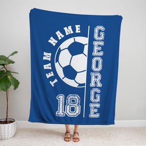 May include: A royal blue blanket with a soccer ball graphic and the words "TEAM NAME" and "18" in white. The name "GEORGE" is printed vertically on the right side. The blanket is displayed indoors.