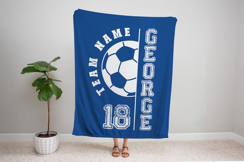 May include: A royal blue blanket with a soccer ball graphic and the words "TEAM NAME" and "18" in white. The name "GEORGE" is printed vertically on the right side. The blanket is displayed indoors.