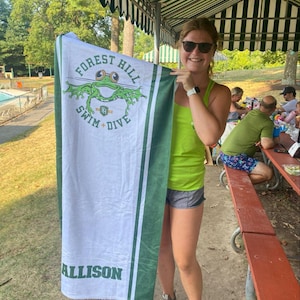 Puede incluir: Toalla de playa blanca y verde con el texto "FOREST HILL SWIM + DIVE" y un dise&ntilde;o de rana. La toalla tiene rayas verdes y el nombre "ALLISON".
