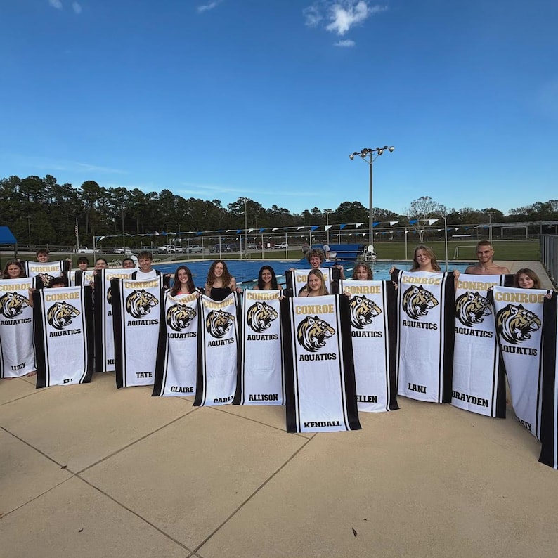 Peut inclure: Un groupe de personnes tenant des serviettes blanches et noires avec le texte "CONROE AQUATICS" et un logo de tigre. Les serviettes ont des bordures noires et sont tenues devant une piscine.