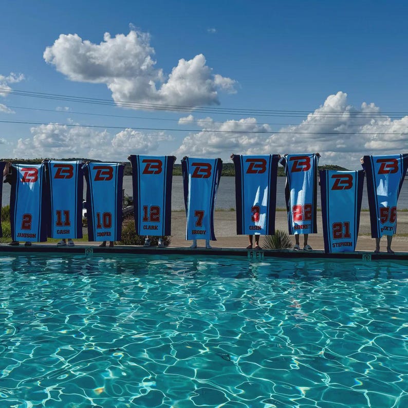 Peut inclure: Rang&eacute;e de serviettes de sport bleues et blanches avec la lettre "B", des num&eacute;ros et des noms imprim&eacute;s. Les serviettes sont tenues par des personnes au bord d'une piscine. Le ciel est bleu avec des nuages blancs.
