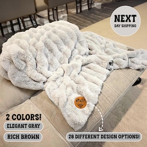 May include: A plush, elegant gray blanket with a textured, rippled design. The blanket is displayed on a beige sofa. The image also shows the text "2 COLORS!" and "28 DIFFERENT DESIGN OPTIONS!"