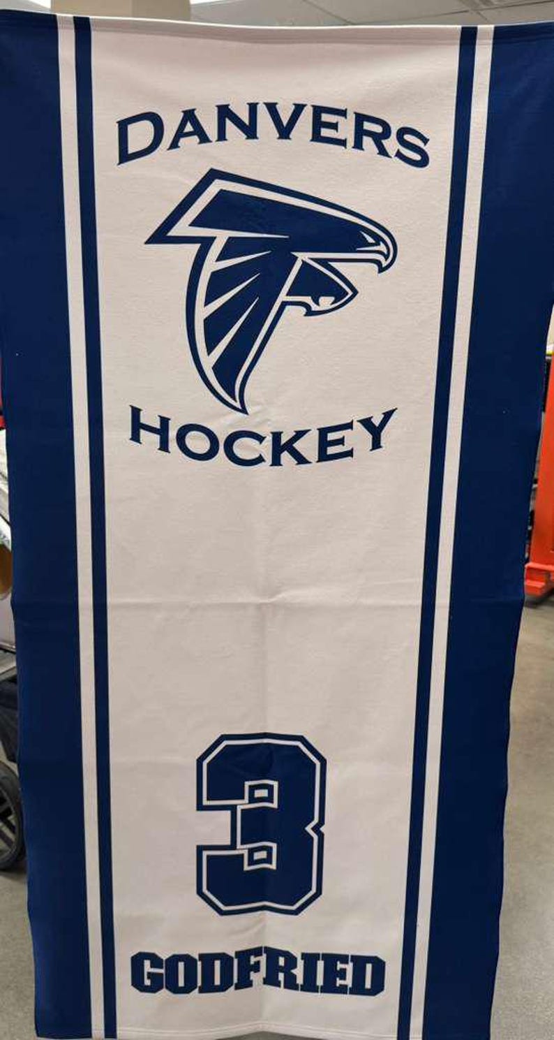 Peut inclure: Serviette de hockey blanche et bleue. Le mot "DANVERS" est en haut, avec un logo de faucon bleu et le mot "HOCKEY" en dessous. Le num&eacute;ro "3" est dans un encadr&eacute; bleu, avec le nom "GODFRIED" en dessous.