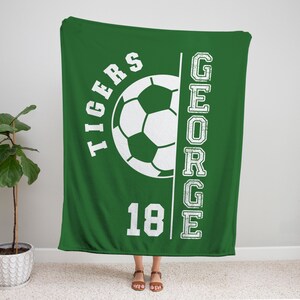 May include: Green blanket with a white soccer ball graphic, the word "TIGERS" above the ball, and the name "GEORGE" vertically on the right. The number "18" is below the ball. The blanket is displayed indoors.