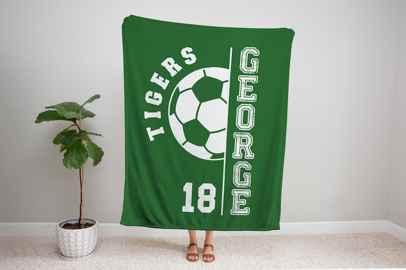 May include: Green blanket with a white soccer ball graphic, the word "TIGERS" above the ball, and the name "GEORGE" vertically on the right. The number "18" is below the ball. The blanket is displayed indoors.