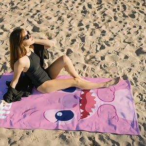 May include: A pink beach towel with a cartoon character's face on it. The character has large eyes, a wide mouth, and a pink nose. A person is lying on the towel wearing sunglasses and a black swimsuit.