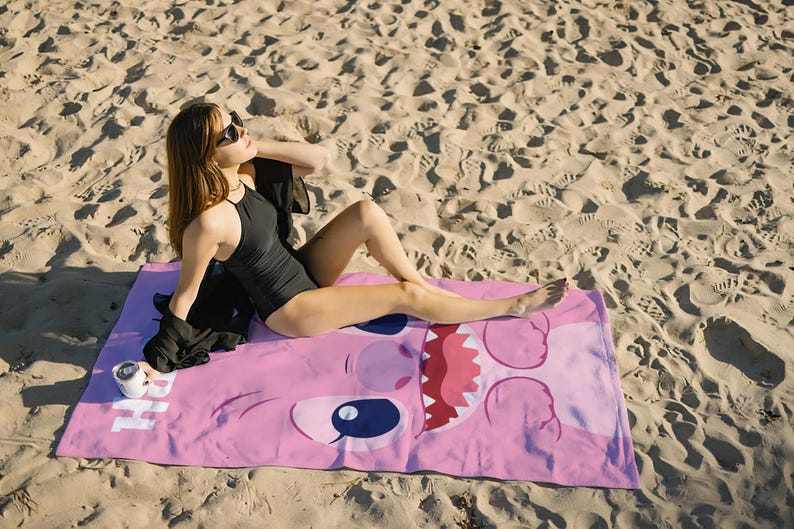 May include: A pink beach towel with a cartoon character's face on it. The character has large eyes, a wide mouth, and a pink nose. A person is lying on the towel wearing sunglasses and a black swimsuit.