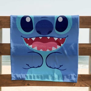 May include: A blue beach towel with a cartoon character of Stitch from Lilo & Stitch. Stitch has big blue eyes, a wide smile, and is shown with his arms crossed.