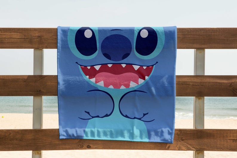 May include: A blue beach towel with a cartoon character of Stitch from Lilo & Stitch. Stitch has big blue eyes, a wide smile, and is shown with his arms crossed.