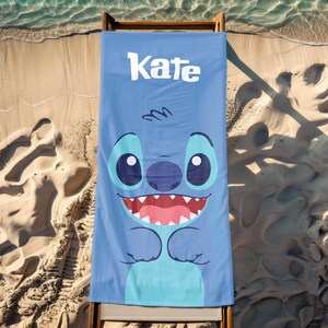 May include: Blue beach towel with a cartoon Stitch character from Lilo & Stitch. The towel has the name "Kate" printed on it in white.