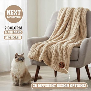 May include: A beige cable-knit throw blanket draped over a gray armchair. The blanket has a soft, textured appearance. The image also shows a cat and text that reads "2 Colors! Warm Sand Arctic Ash" and "28 Different Design Options!"