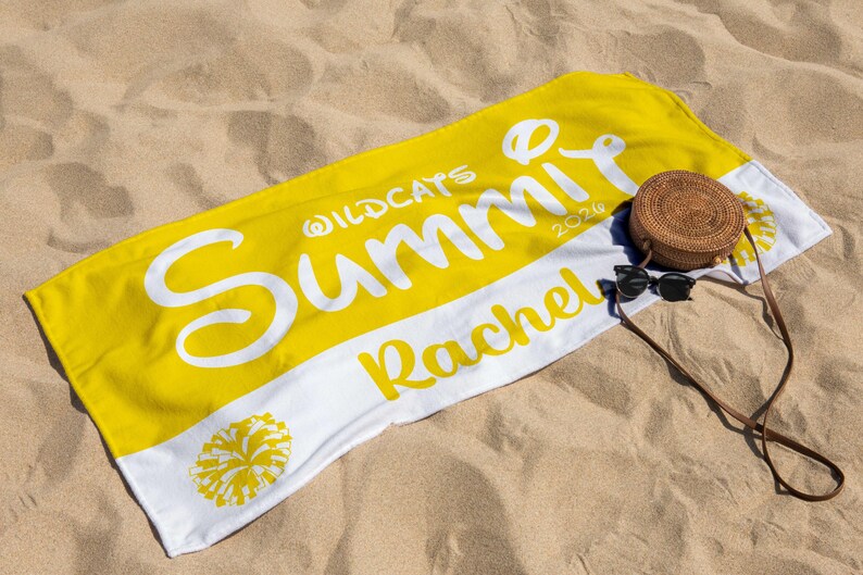 May include: Yellow and white beach towel with the text "Wildcats Summit 2026 Rachel". A round woven bag and sunglasses rest on the towel, which is on a sandy surface.