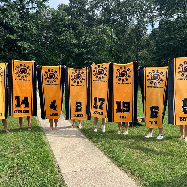 Peut inclure: Plusieurs serviettes de sport dor&eacute;es et noires avec le texte "Gold Coast Hurricanes" et des num&eacute;ros de joueurs. Chaque serviette affiche un num&eacute;ro et un nom de joueur diff&eacute;rents, dont 14 Chelsea, 4 Reese, 2 Reese, 17 Karlie, 19 Lily et 6 Tata.