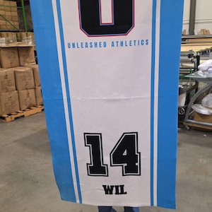 Puede incluir: Bandera rectangular con centro blanco y bordes azul claro. La bandera presenta una gran "U" negra y roja y el texto "UNLEASHED ATHLETICS". Debajo, el n&uacute;mero "14" en blanco y negro, con el nombre "WIL" debajo.