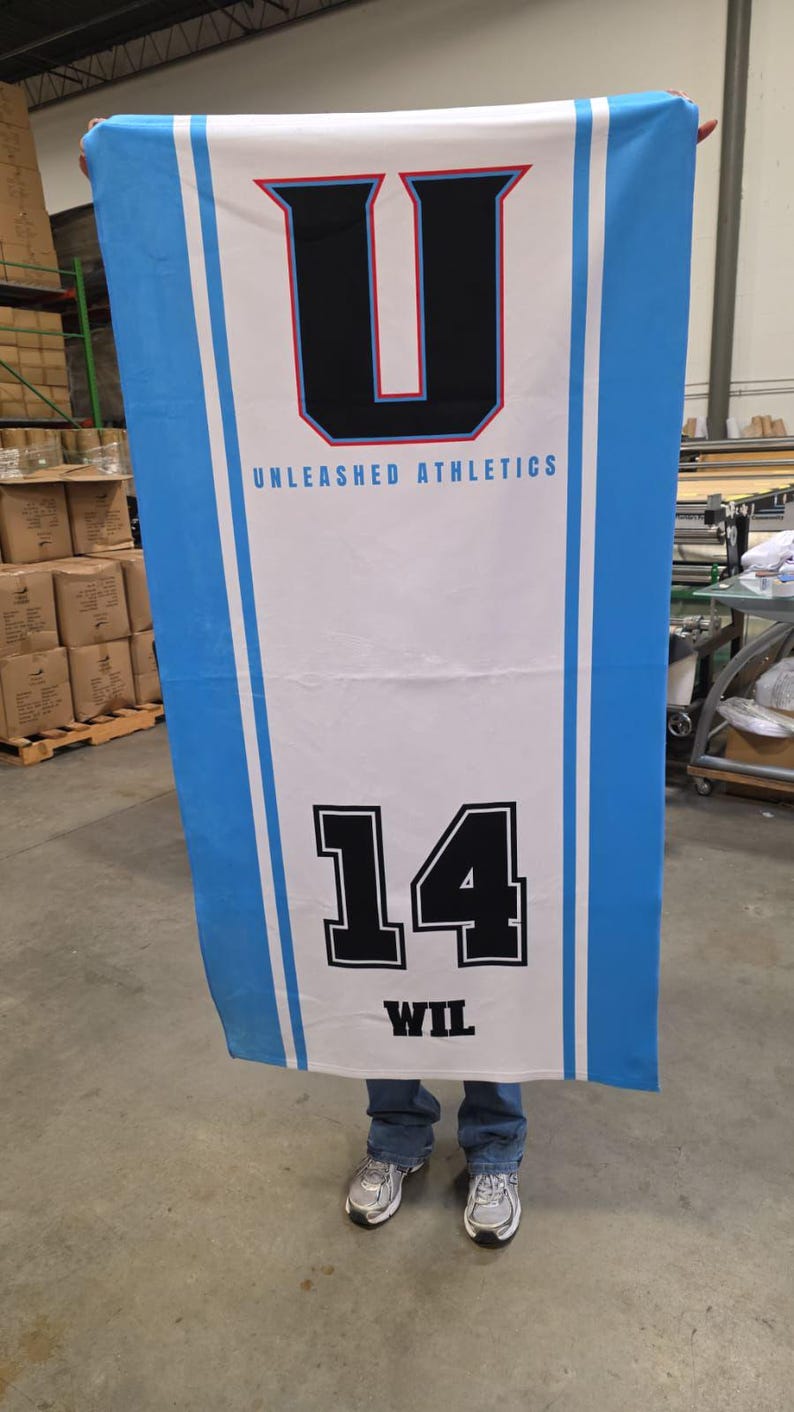 Puede incluir: Bandera rectangular con centro blanco y bordes azul claro. La bandera presenta una gran "U" negra y roja y el texto "UNLEASHED ATHLETICS". Debajo, el n&uacute;mero "14" en blanco y negro, con el nombre "WIL" debajo.