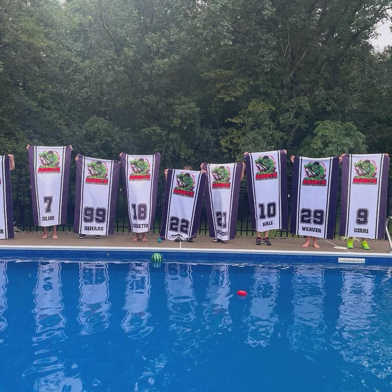 Peut inclure: Plusieurs personnes tiennent des serviettes blanches et violettes avec des num&eacute;ros et des logos d'&eacute;quipe. Chaque serviette affiche un grand num&eacute;ro et un logo d'&eacute;quipe. Les serviettes sont devant une piscine.