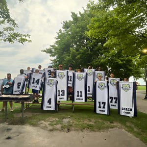 Puede incluir: Un grupo de personas sosteniendo camisetas de f&uacute;tbol con n&uacute;meros y nombres. Las camisetas son blancas con detalles en azul marino y un logotipo del equipo. Los n&uacute;meros van del 1 al 77. Tambi&eacute;n se ve la palabra "COACH".