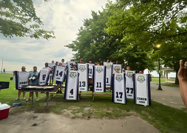 Puede incluir: Un grupo de personas sosteniendo camisetas de f&uacute;tbol con n&uacute;meros y nombres. Las camisetas son blancas con detalles en azul marino y un logotipo del equipo. Los n&uacute;meros van del 1 al 77. Tambi&eacute;n se ve la palabra "COACH".
